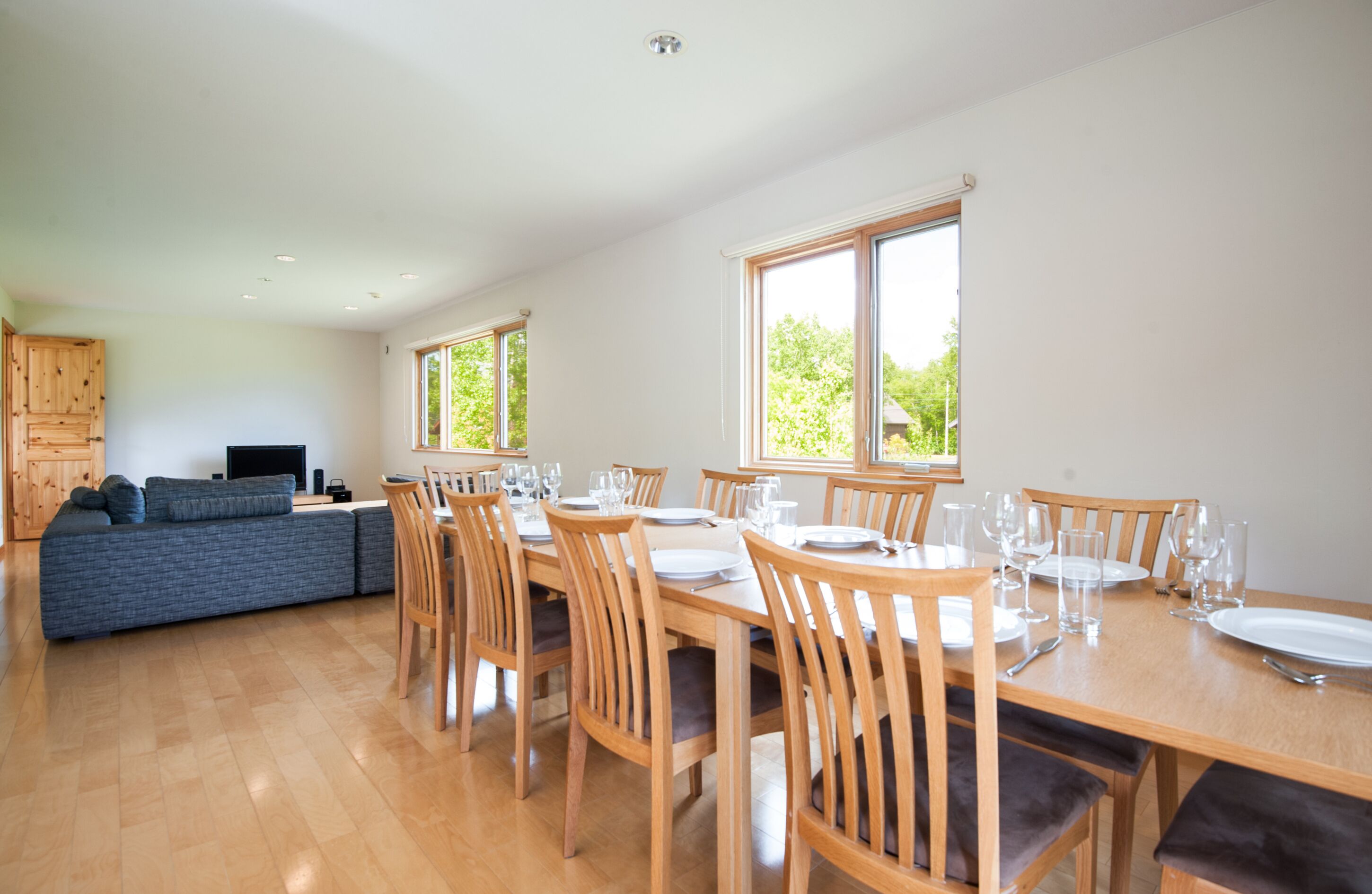 A dining area with a wooden table set for six, surrounded by six chairs. There is a blue sofa in the background, and a television is mounted on the wall.