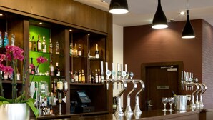 A bar area with a counter, multiple taps, a cash register, and a green display. There are bar stools and a potted plant with pink flowers. - Leonardo Hotel Derby (Derby)