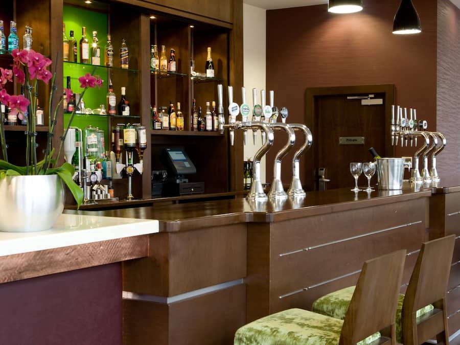 A bar area with a counter, multiple taps, a cash register, and a green display. There are bar stools and a potted plant with pink flowers.