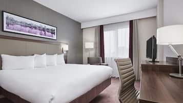 A hotel room with a large bed, a desk with a flat-screen TV, a chair, a desk lamp, and a wall-mounted TV.