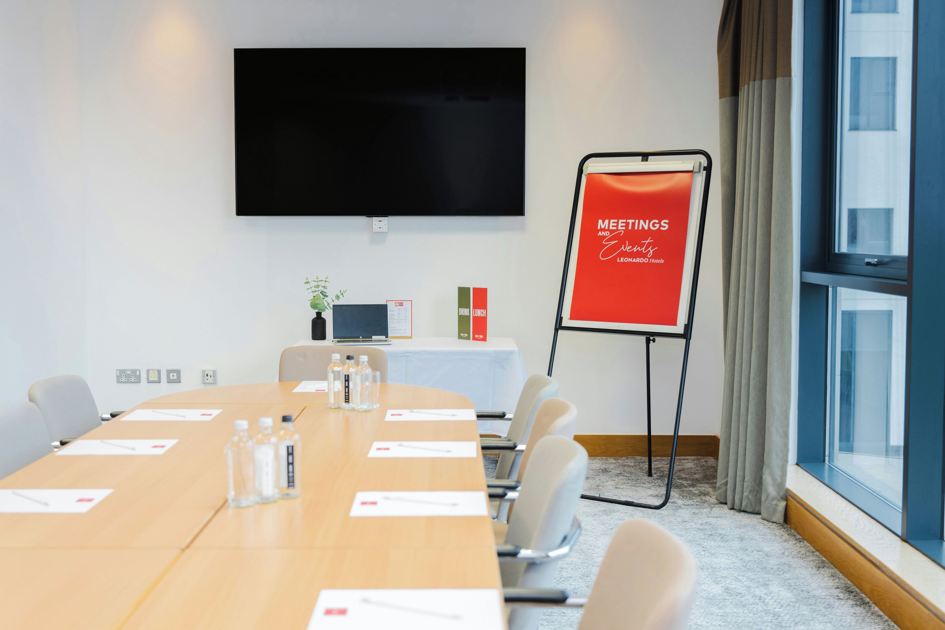 A conference room with a long table, chairs, a TV screen, a standee, and a window with curtains.