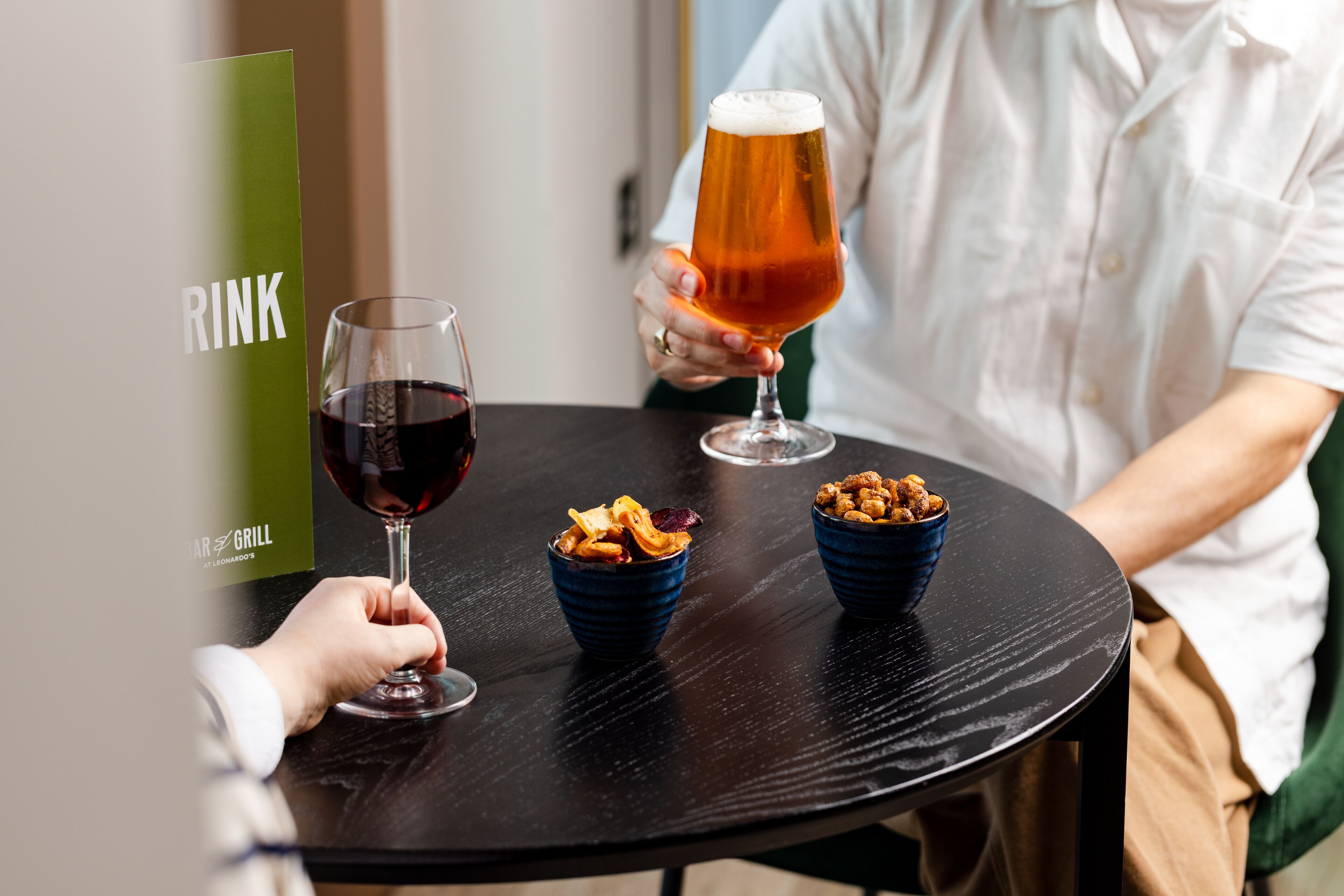 A person holding a glass of beer at a table with two bowls of snacks.