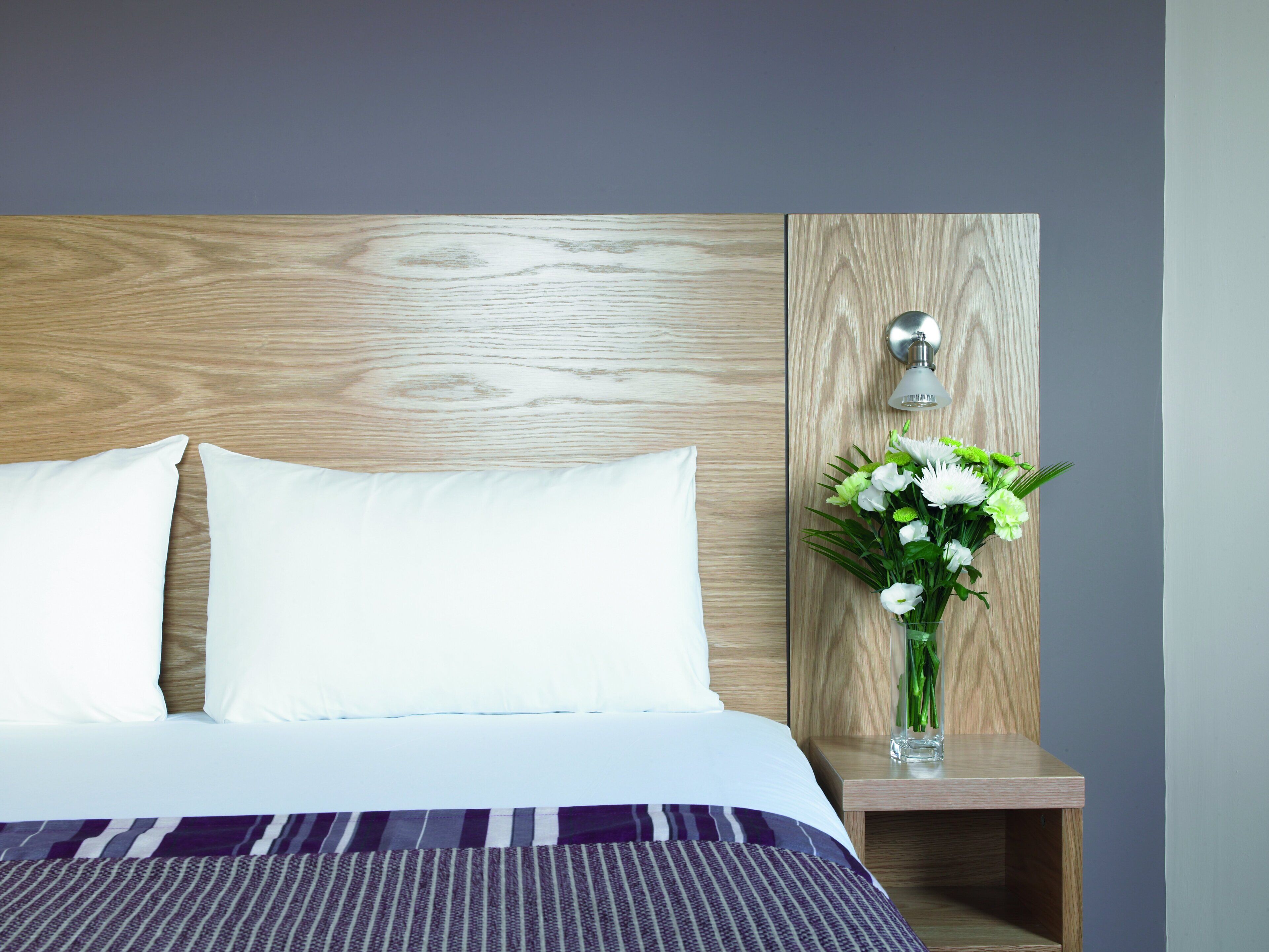 A neatly made bed with white pillows and a striped blanket, a wooden headboard, a bedside table with a vase of flowers, and a wall-mounted lamp.