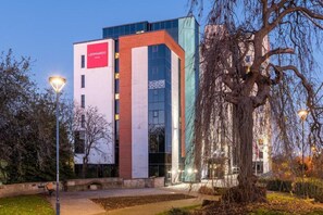 A modern building with a prominent red and white sign, a glass facade, and a tree in the foreground. - Leonardo Hotel Derby (Derby)