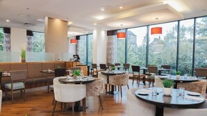 A modern restaurant with round tables and cushioned chairs, large windows with a view of greenery, and a bar area with a counter and seating. - Leonardo Hotel Derby (Derby)