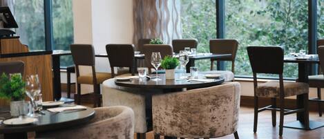 L’intérieur d’un restaurant avec des tables rondes, des chaises et une vue sur la verdure grâce à de grandes fenêtres.