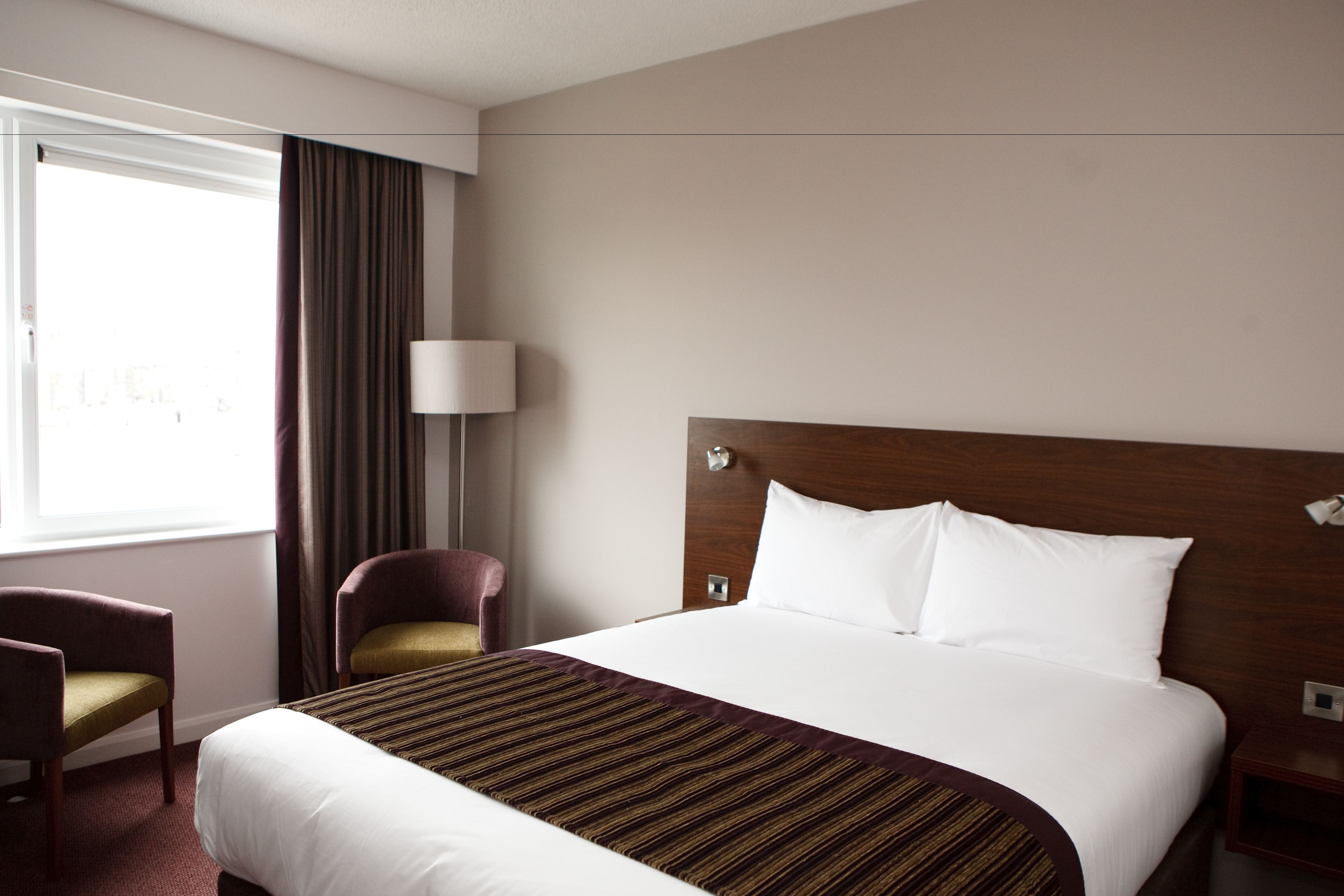 A hotel room with a large bed, a chair, a lamp, and a window with curtains.