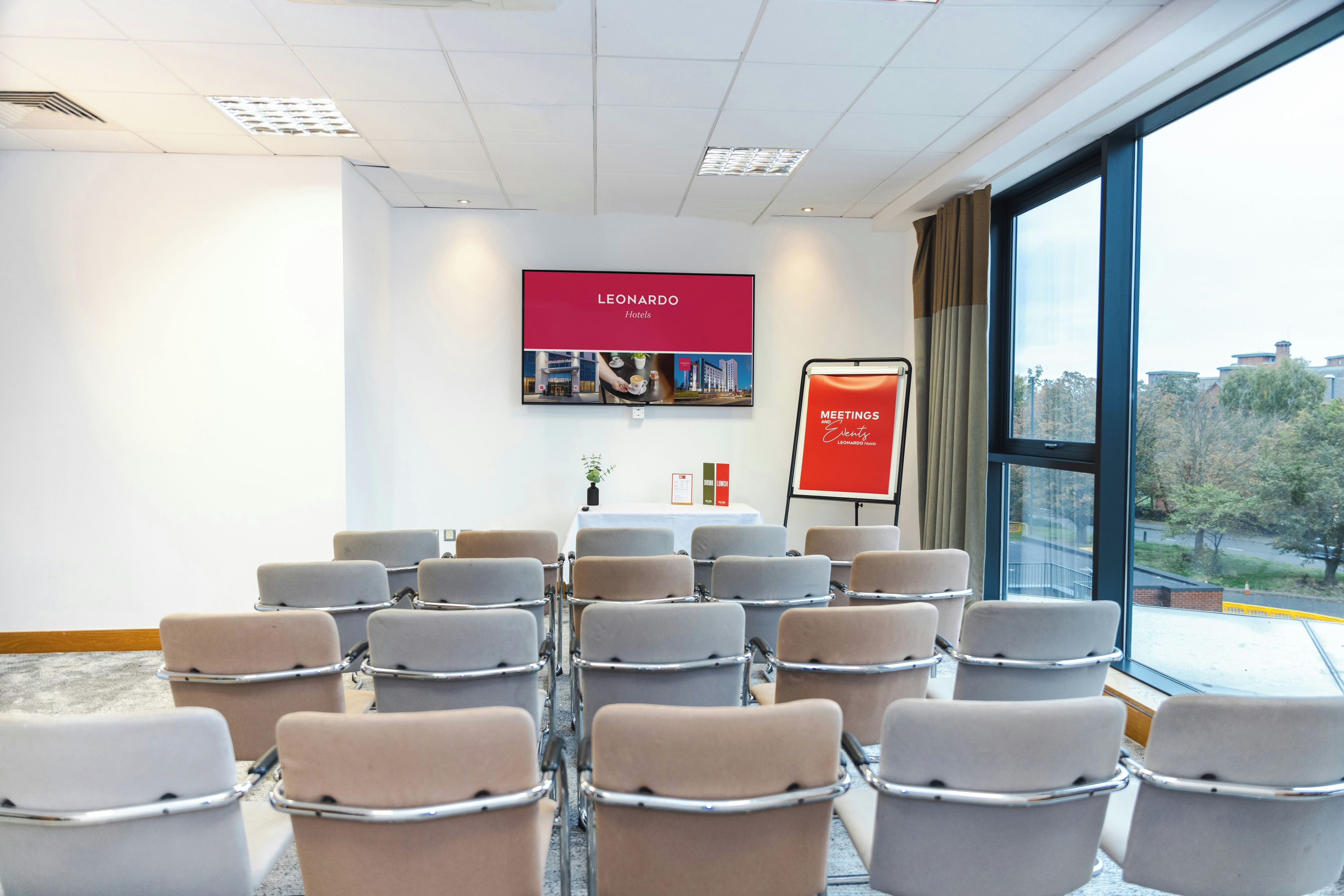 A conference room with rows of chairs, a projector screen displaying hotel information, and a window with a view of buildings and trees.