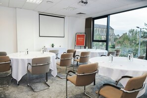 A conference room with round tables, chairs, a projector screen, and a view of a building through large windows. - Leonardo Hotel Derby (Derby)