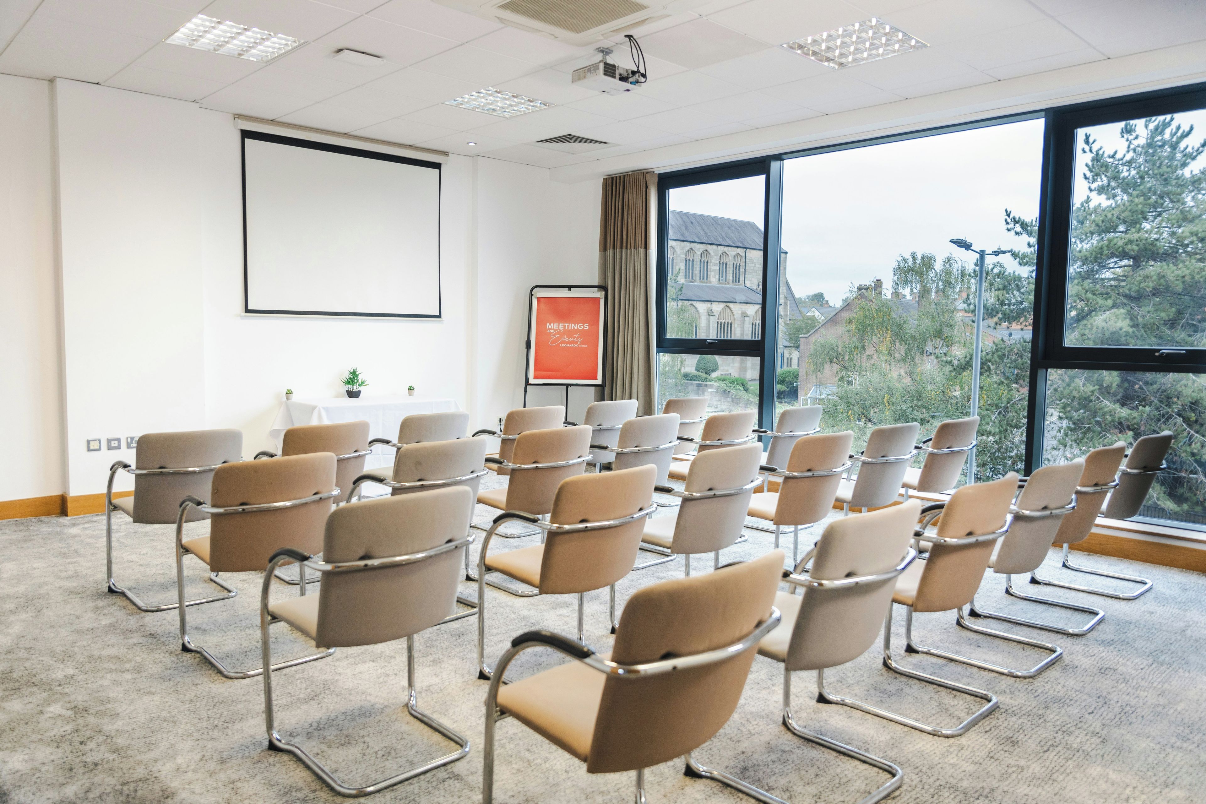 Sala de conferências com fileiras de cadeiras, uma tela de projetor e um quadro de apresentações.