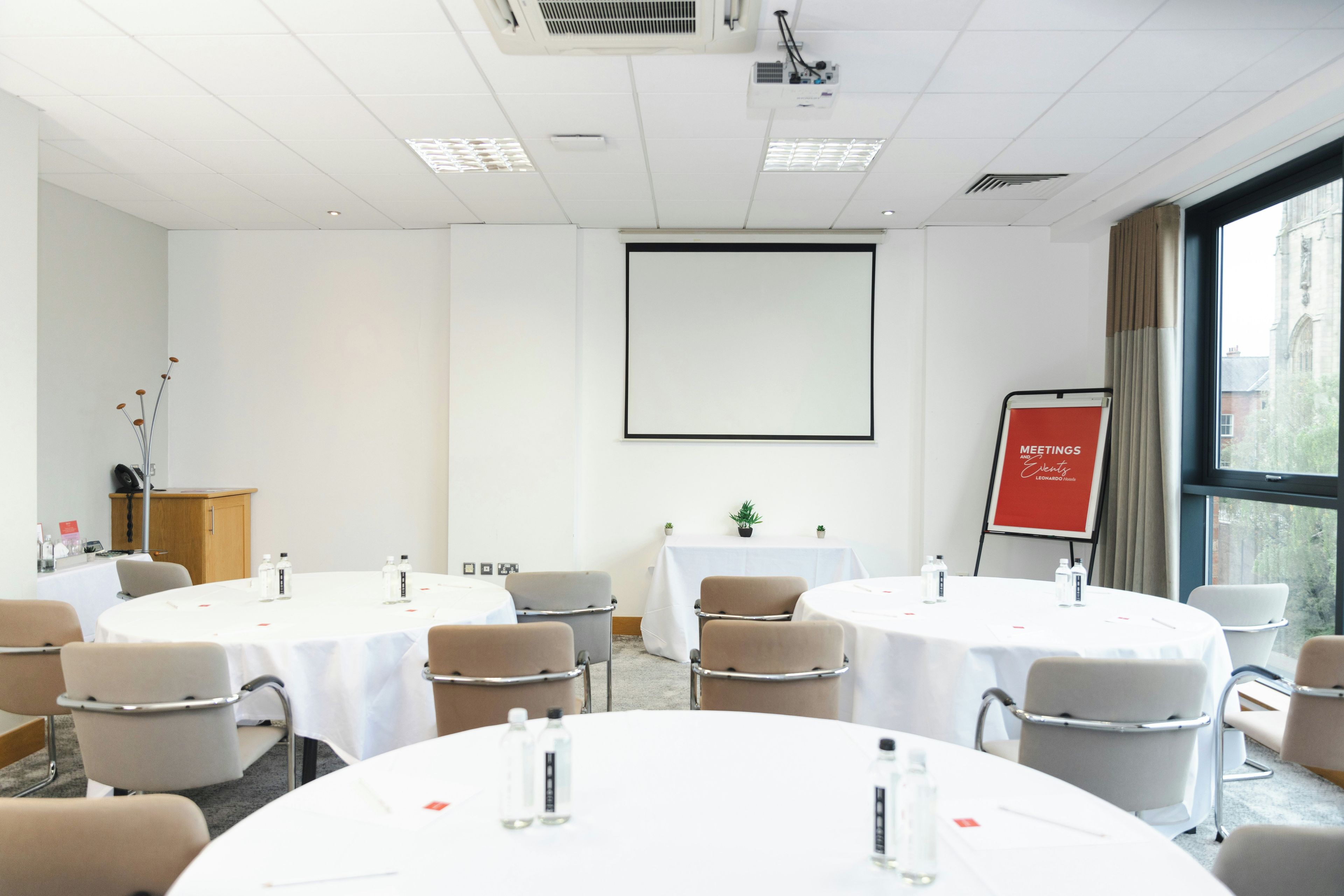 A conference room with round tables, chairs, a projector screen, and a red sign indicating meetings and events.