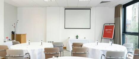A conference room with round tables, chairs, a projector screen, and a red sign indicating meetings and events.