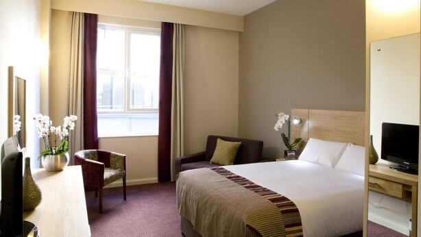 A hotel room with a bed, a chair, a desk with a computer, a television, and a window with curtains.