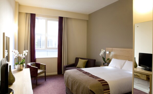 A hotel room with a bed, a chair, a desk with a computer, a television, and a window with curtains.