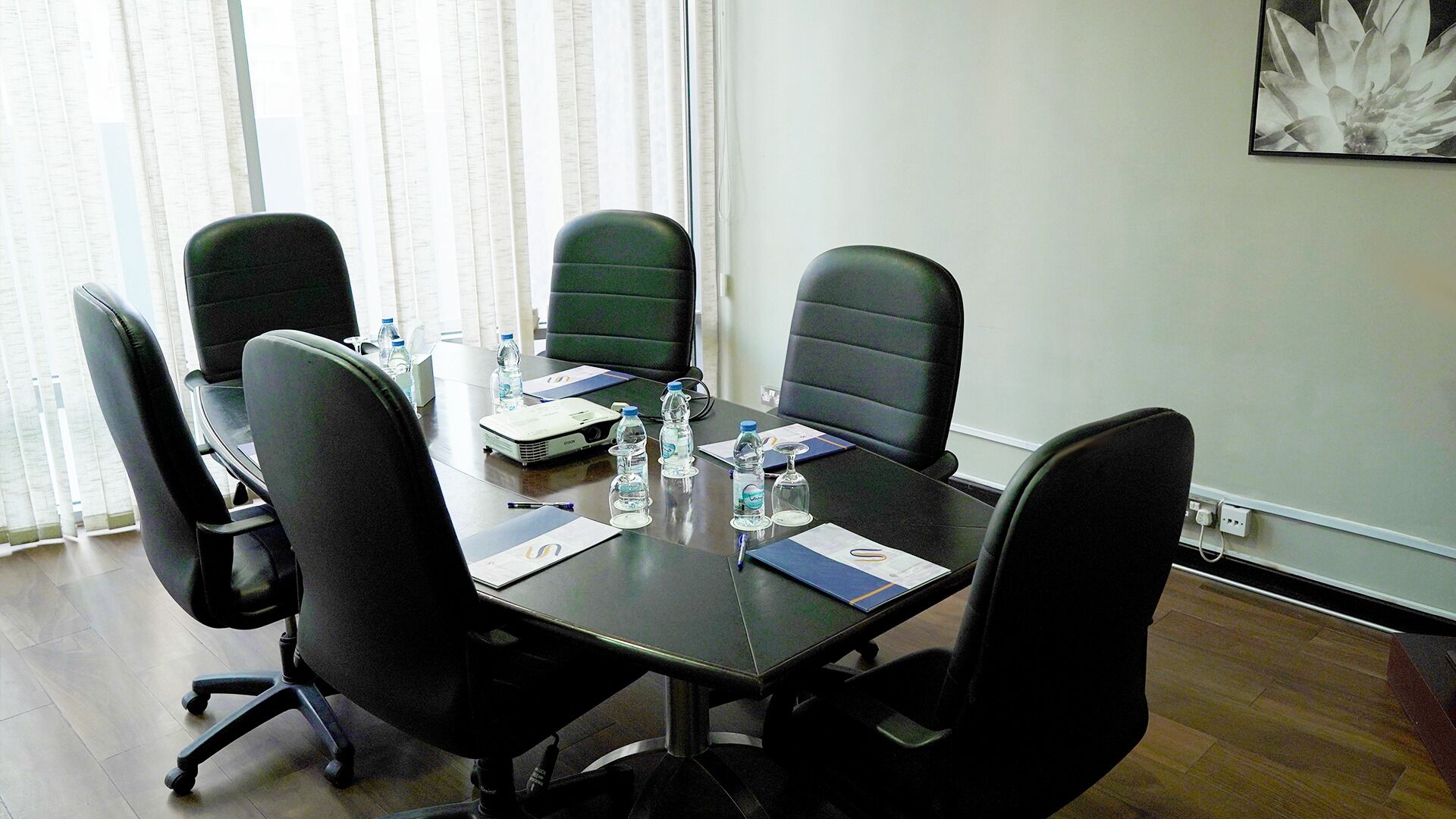 A conference room with a dark table, four black chairs, water bottles, a projector, and a framed picture on the wall.