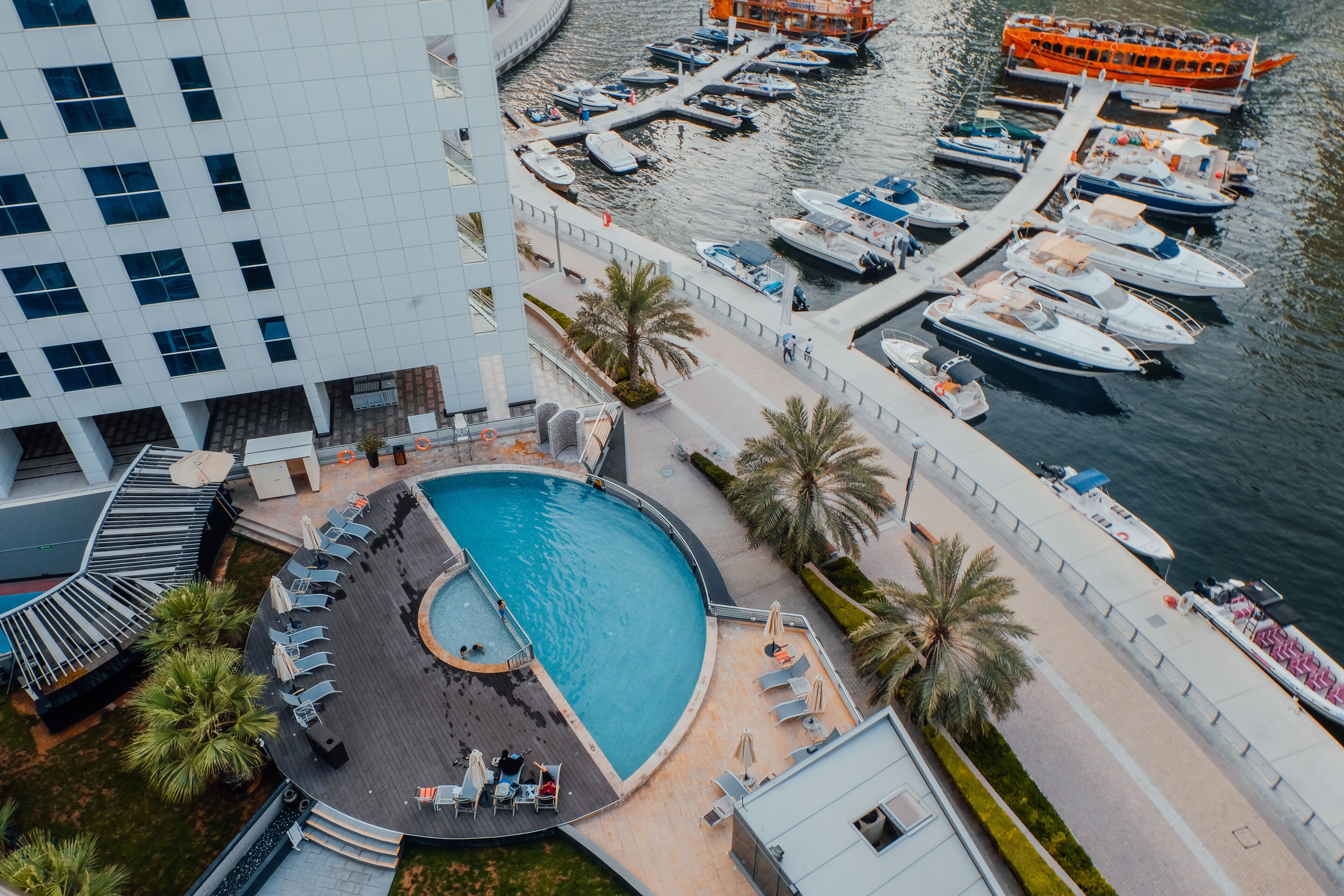 An aerial view of a hotel with a large swimming pool, a marina with boats, and a modern building with a unique architectural design.