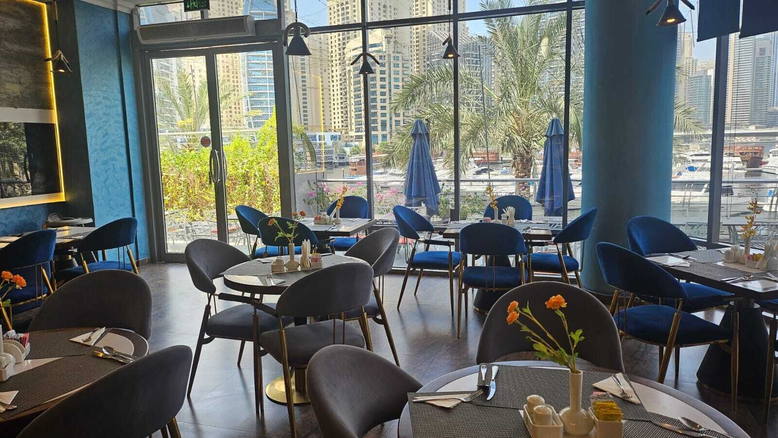 Un restaurant avec des tables rondes et des chaises bleues, de grandes fenêtres offrant une vue sur de hauts immeubles et des palmiers.