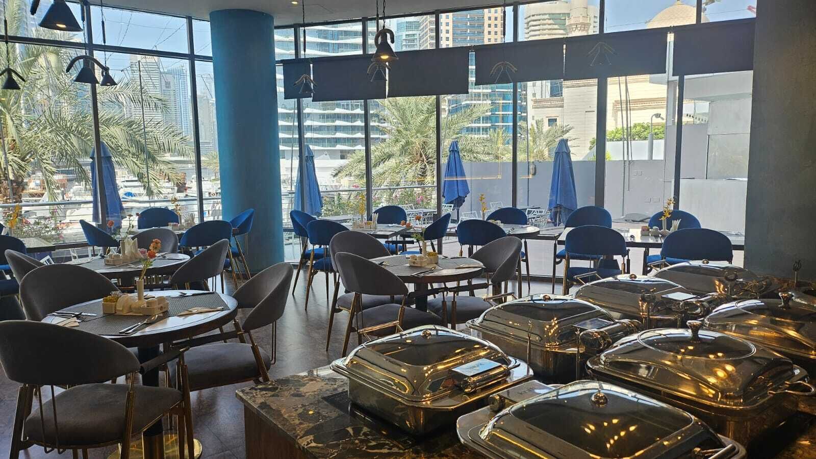 Un restaurant doté de tables et de chaises disposées pour accueillir les clients, de grandes fenêtres offrant une vue sur le paysage urbain et d’un décor intérieur moderne.