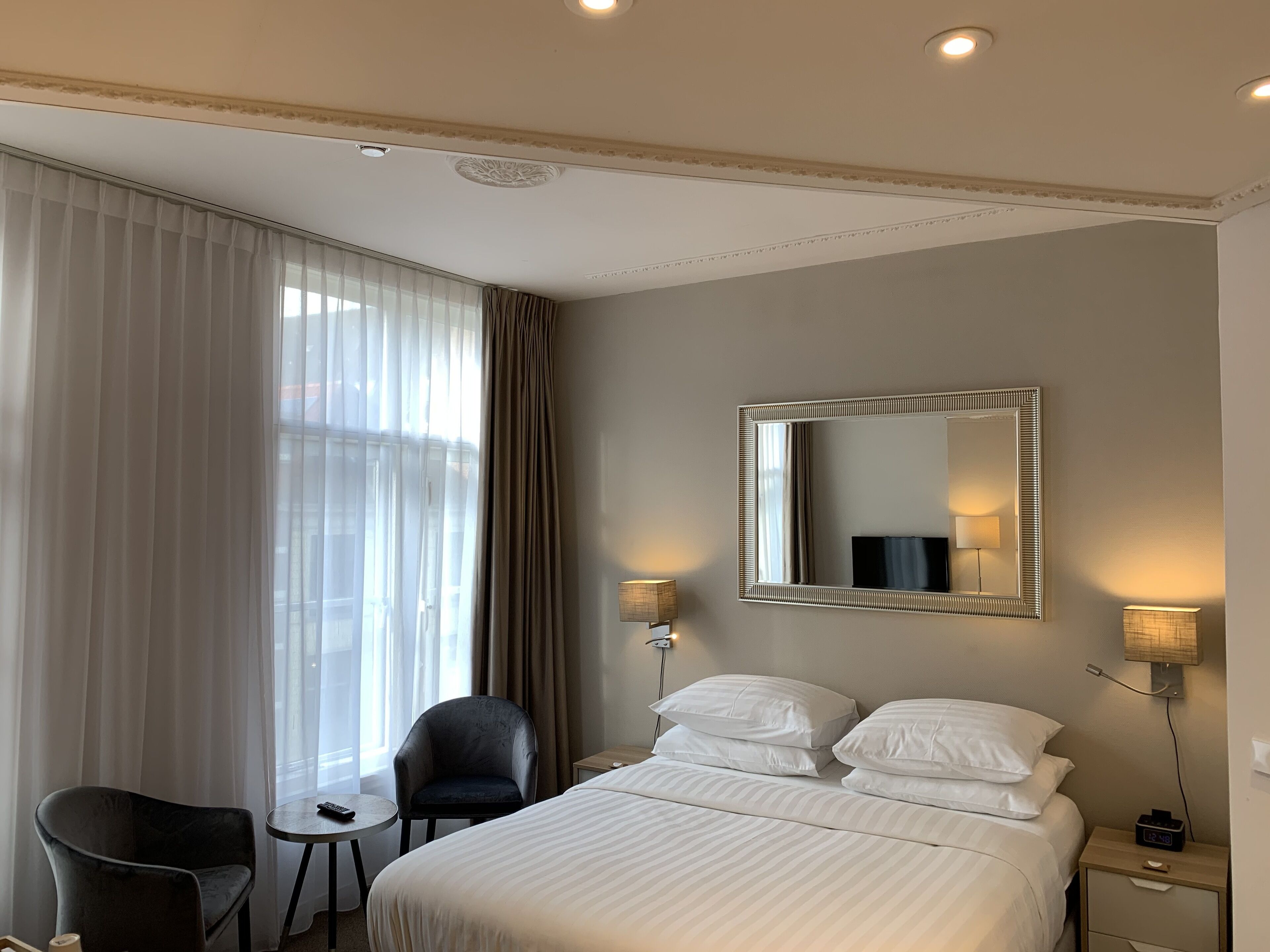 A hotel room with a bed, a chair, a small table, a nightstand with a lamp, and a large mirror on the wall.