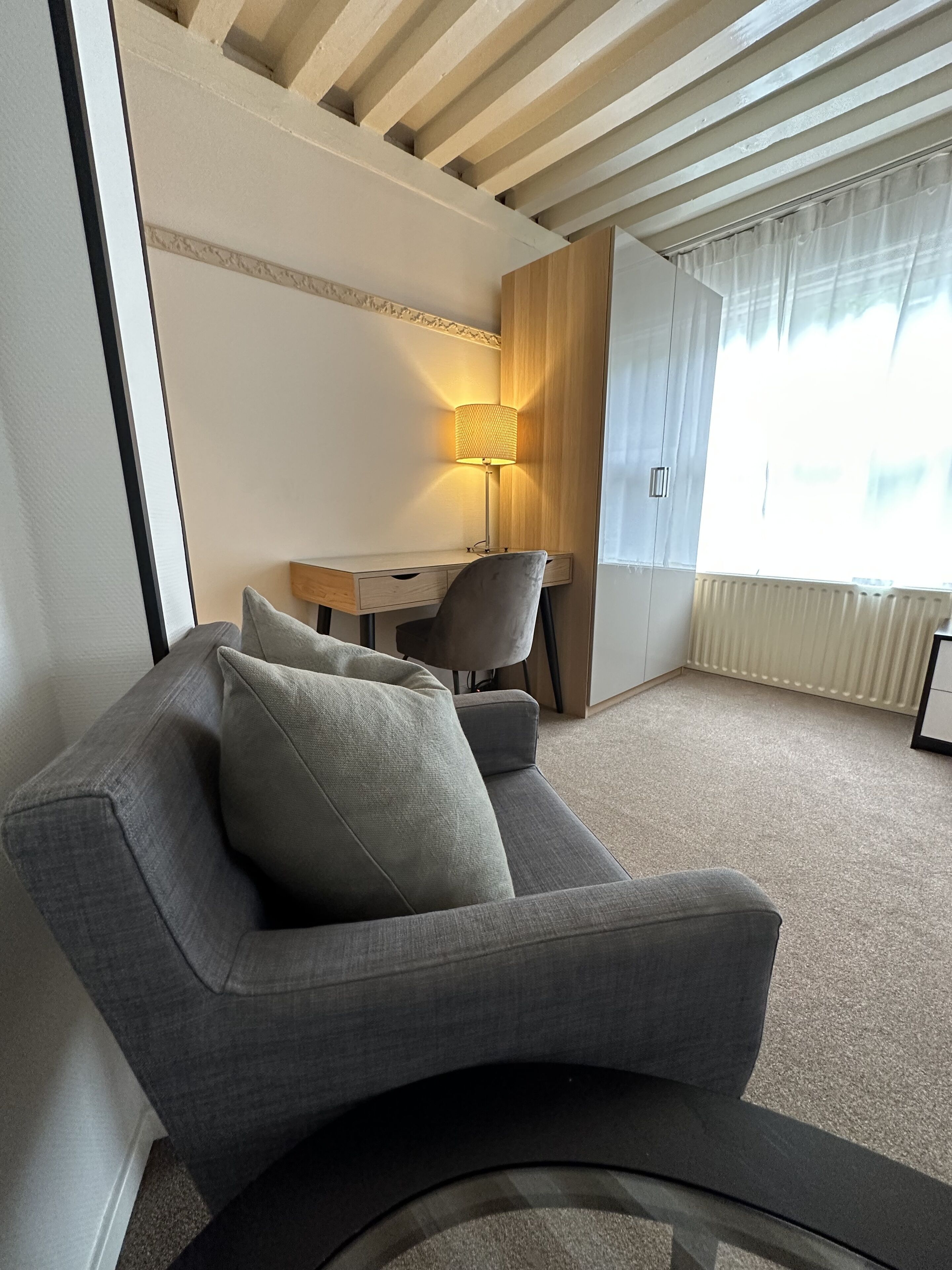 Een moderne hotelkamer met een grijze bank, een houten bureau en een lamp.