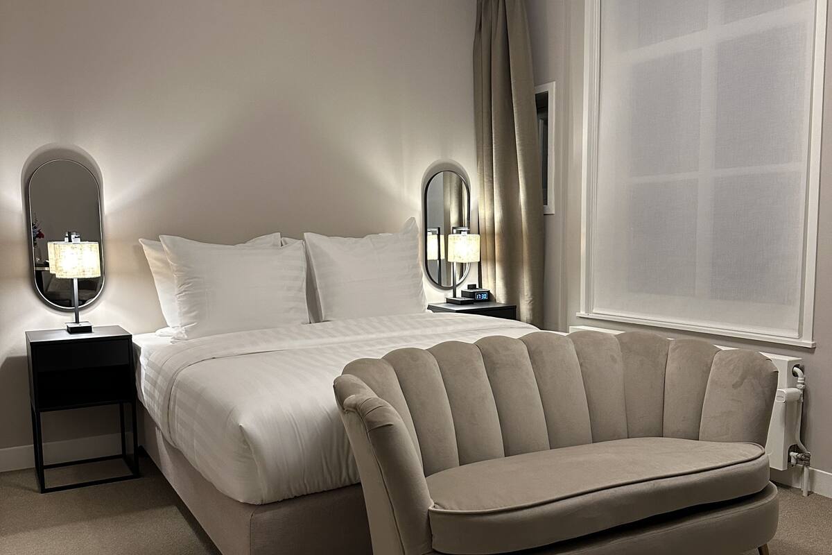 A modern bedroom with a large bed, two bedside tables with lamps, a round mirror, and a sofa.