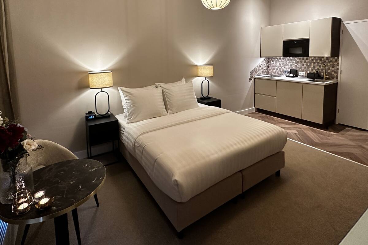 A modern hotel room with a large bed, bedside lamps, a small table with candles, and a kitchenette.