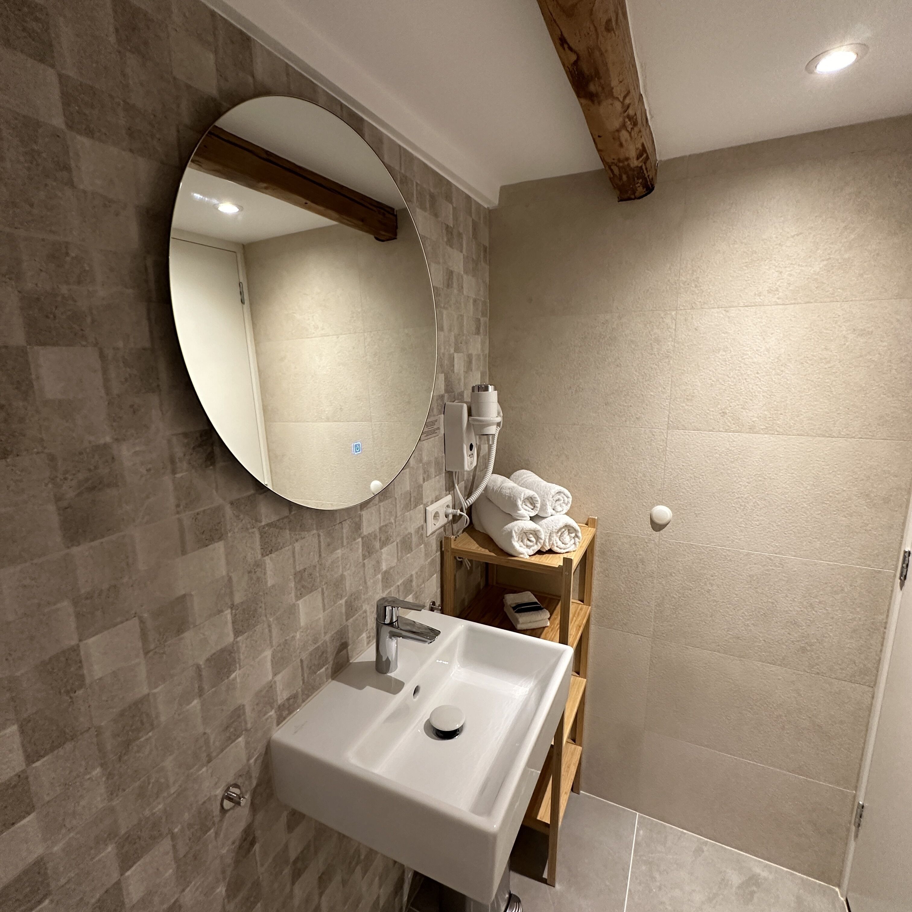 Un baño con un lavamanos blanco, un espejo redondo y un estante de madera con toallas.