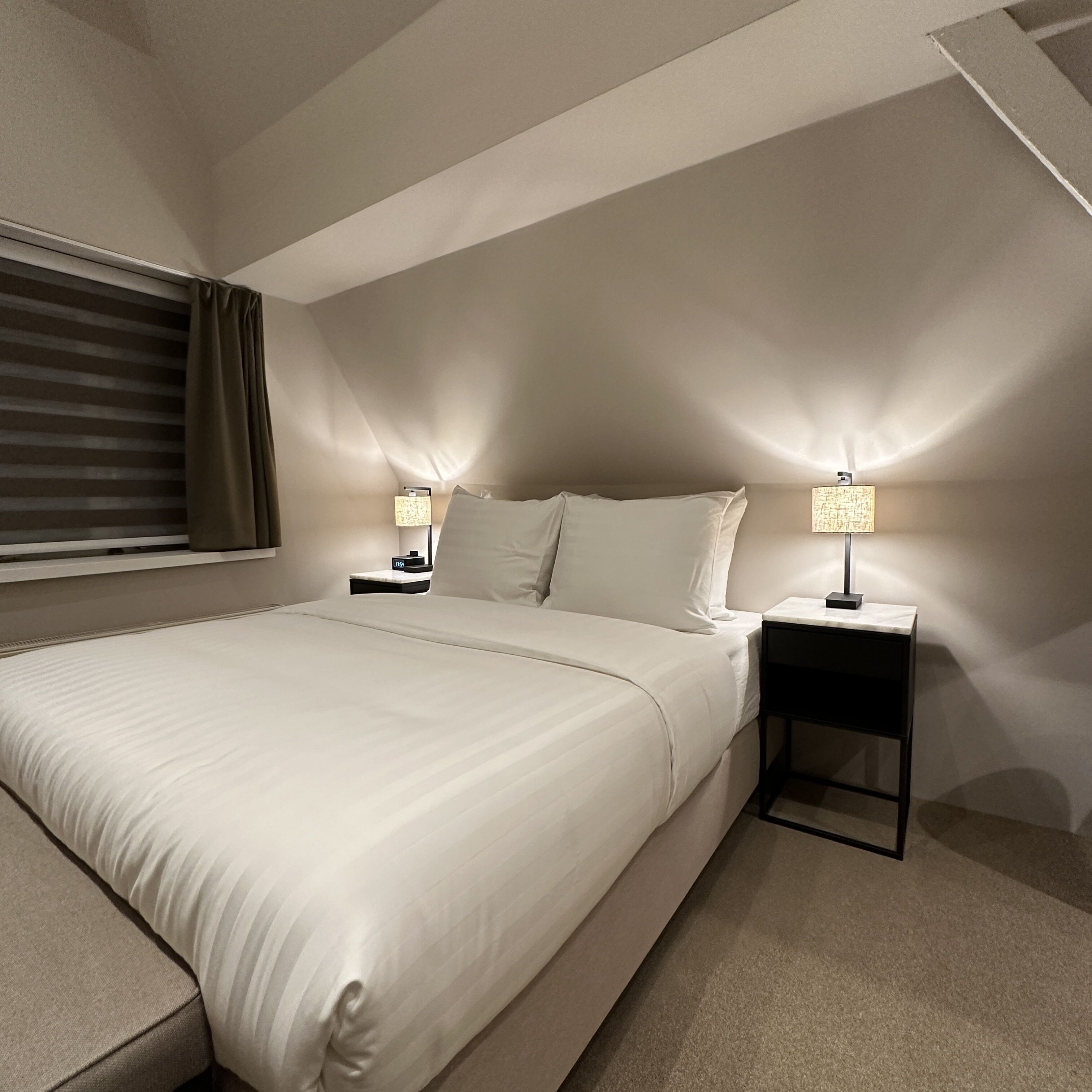 A modern hotel room with a large bed, two bedside tables with lamps, a window with blinds, and a carpeted floor.