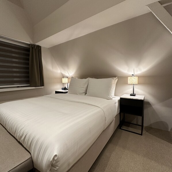 Una habitación de hotel moderna con una cama grande, dos mesitas de noche con lámparas, una ventana con persianas y suelo de moqueta.