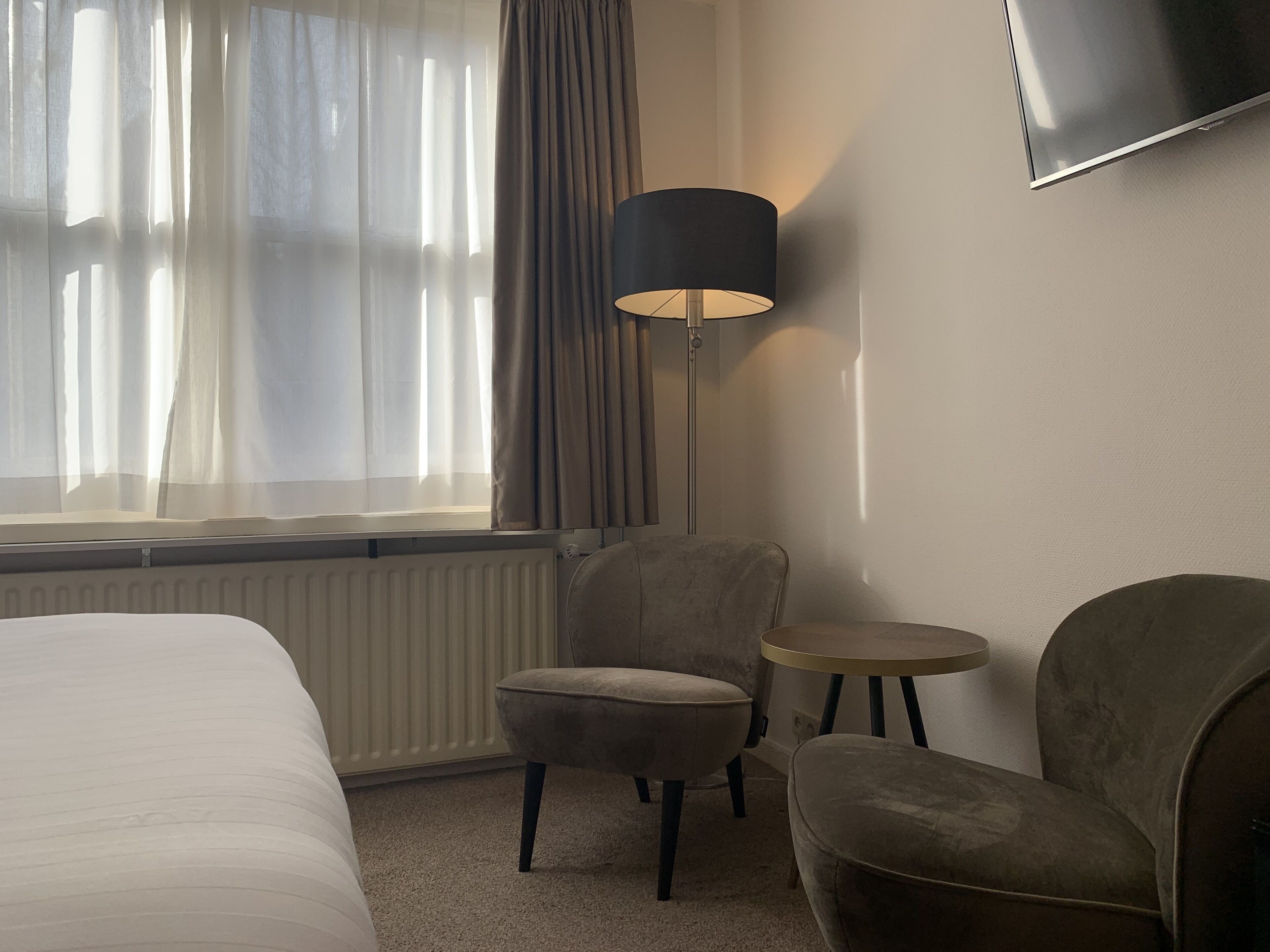 A hotel room with a bed, a chair, a small round table, a lamp, and a TV on the wall.