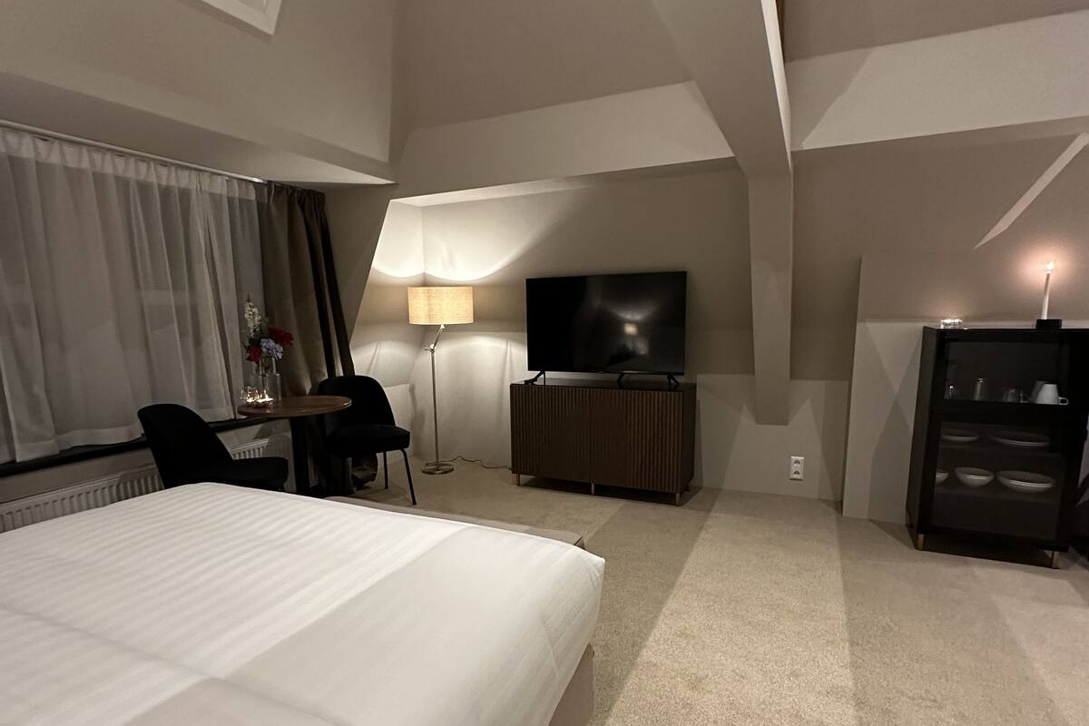 A hotel room with a large bed, a desk, a chair, a TV, a lamp, and a cabinet.