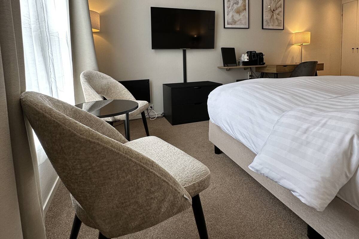 A hotel room with a bed, a chair, a desk, a TV, and artwork on the wall.