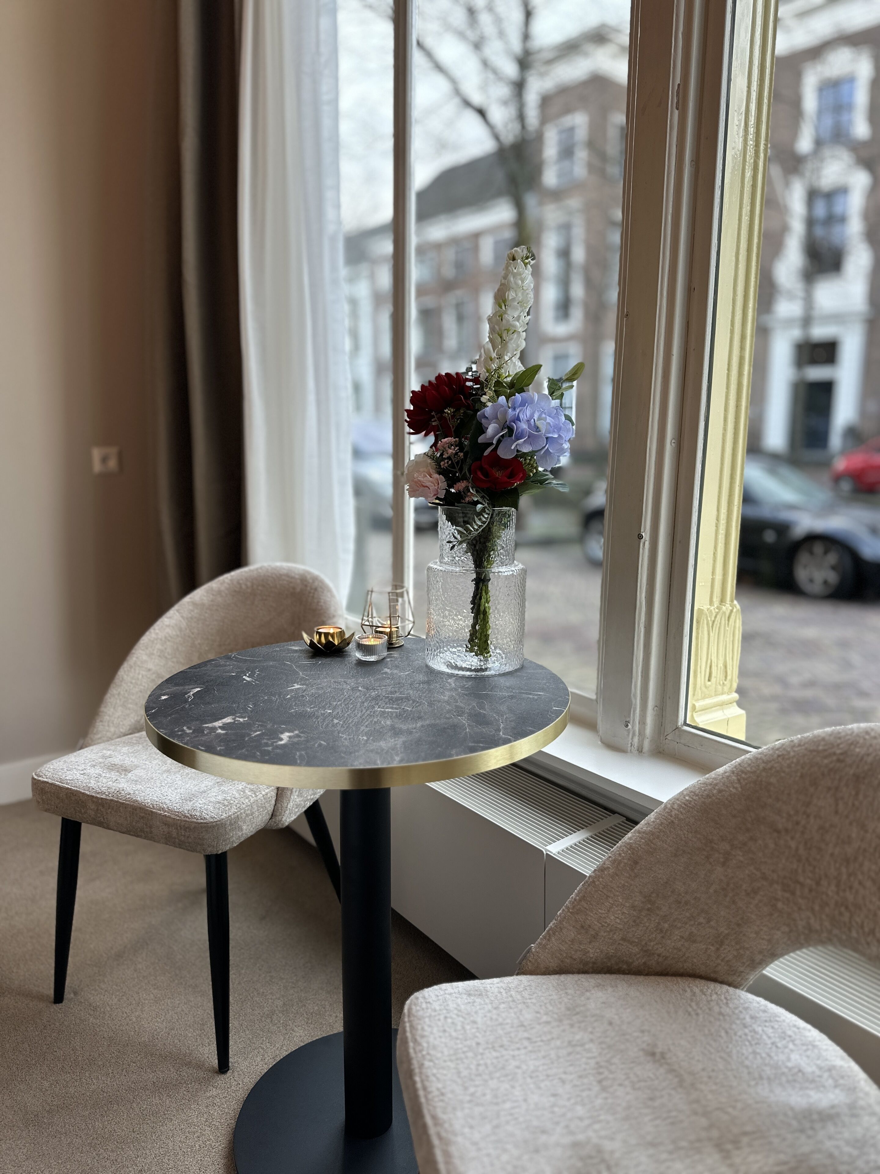 Una mesa redonda con tapa de mármol y patas negras, acompañada por dos sillas color beige. Un florero de vidrio con flores está colocado sobre la mesa.