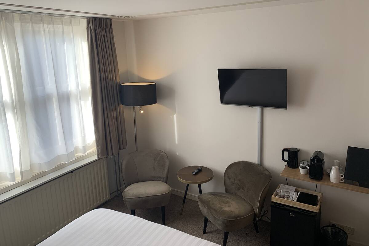 A hotel room with a bed, two armchairs, a small table, a TV, a lamp, and a coffee station.