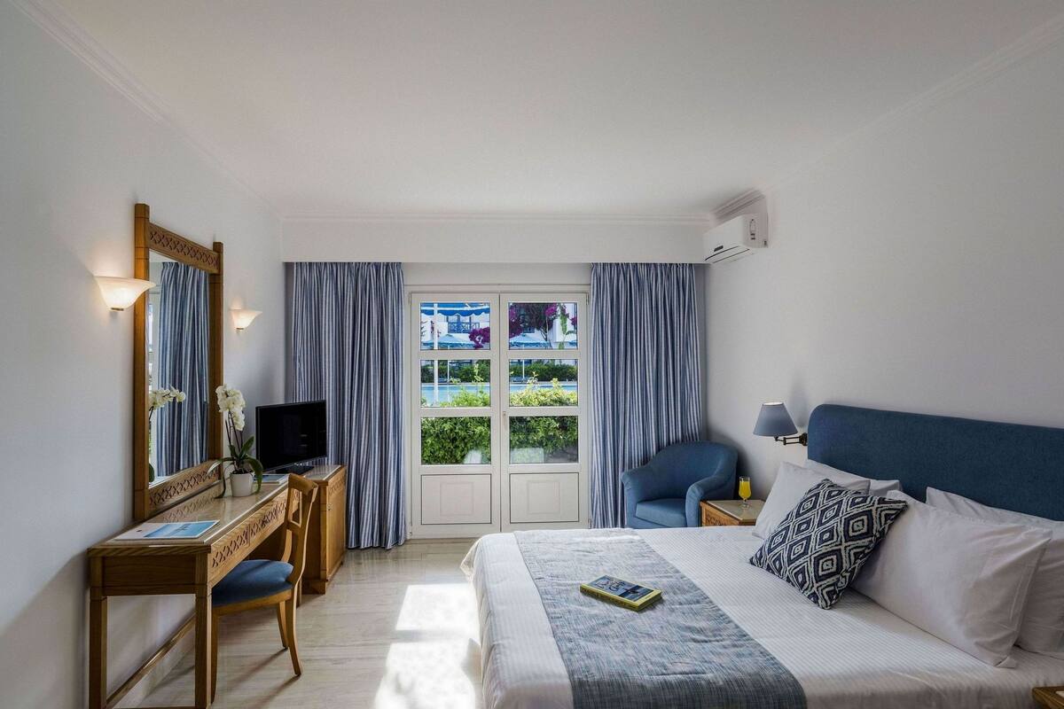 A hotel room with a bed, a desk with a computer, a chair, a TV, and a window with curtains.