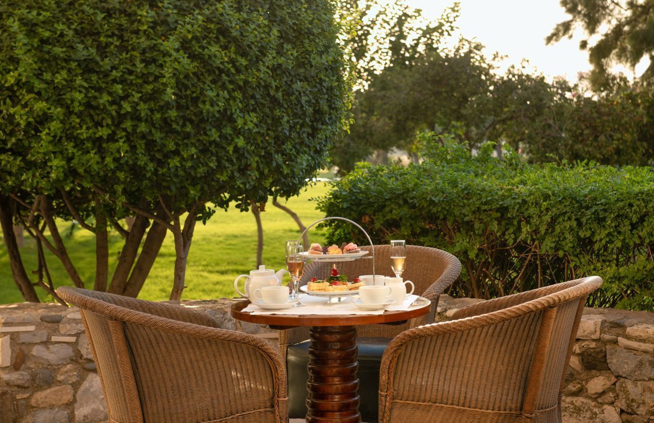A wicker table with a tea set and a dessert tray, surrounded by chairs, set against a stone wall and lush greenery.
