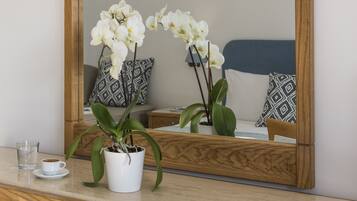 A wooden desk with a white orchid in a white pot, a glass of water, and a cup of coffee. A large mirror reflects a bed with patterned pillows.