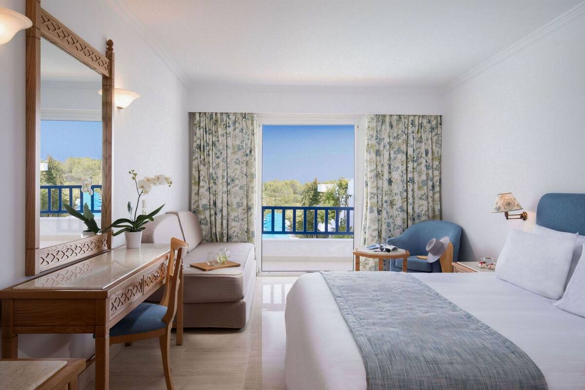 A hotel room with a large bed, a desk, a chair, a small table, and a balcony with a view.