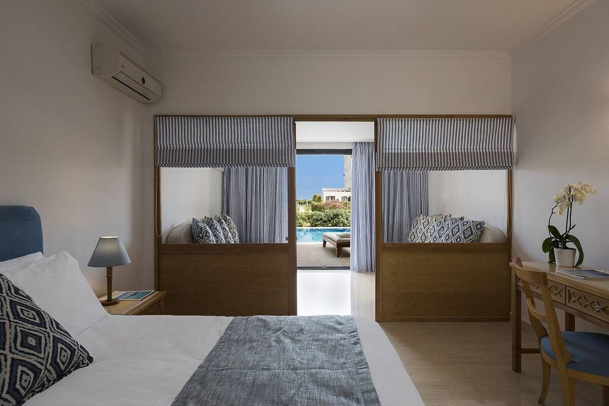 A hotel room with a bed, a desk, a chair, a lamp, and a view of a pool and greenery.