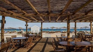 Een terras met houten tafels en stoelen, met uitzicht op een strand met parasols en ligbedden.