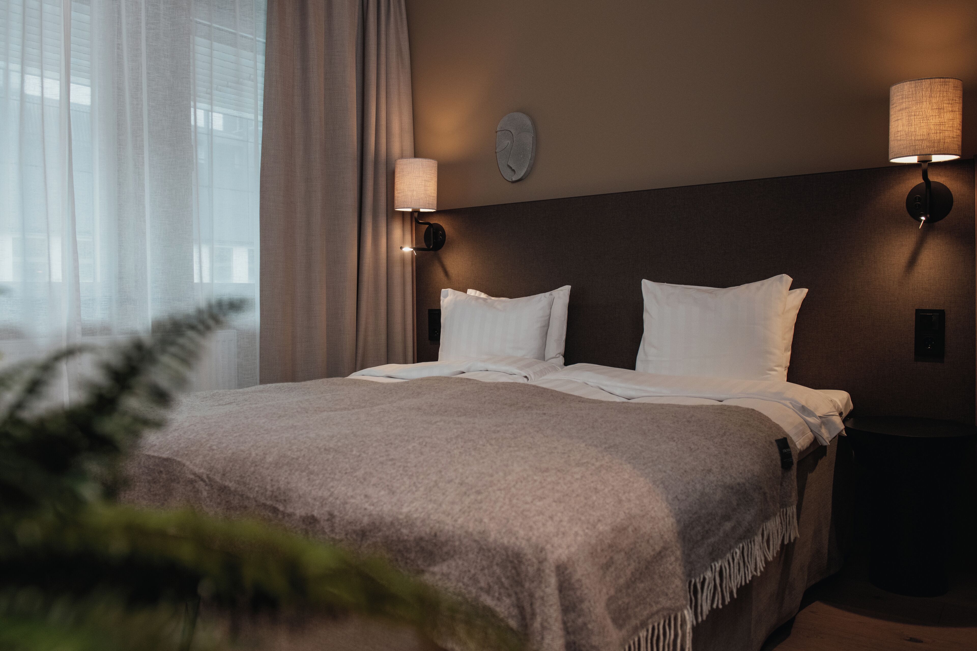 A hotel room with a large bed, two wall-mounted lamps, and a window with curtains.