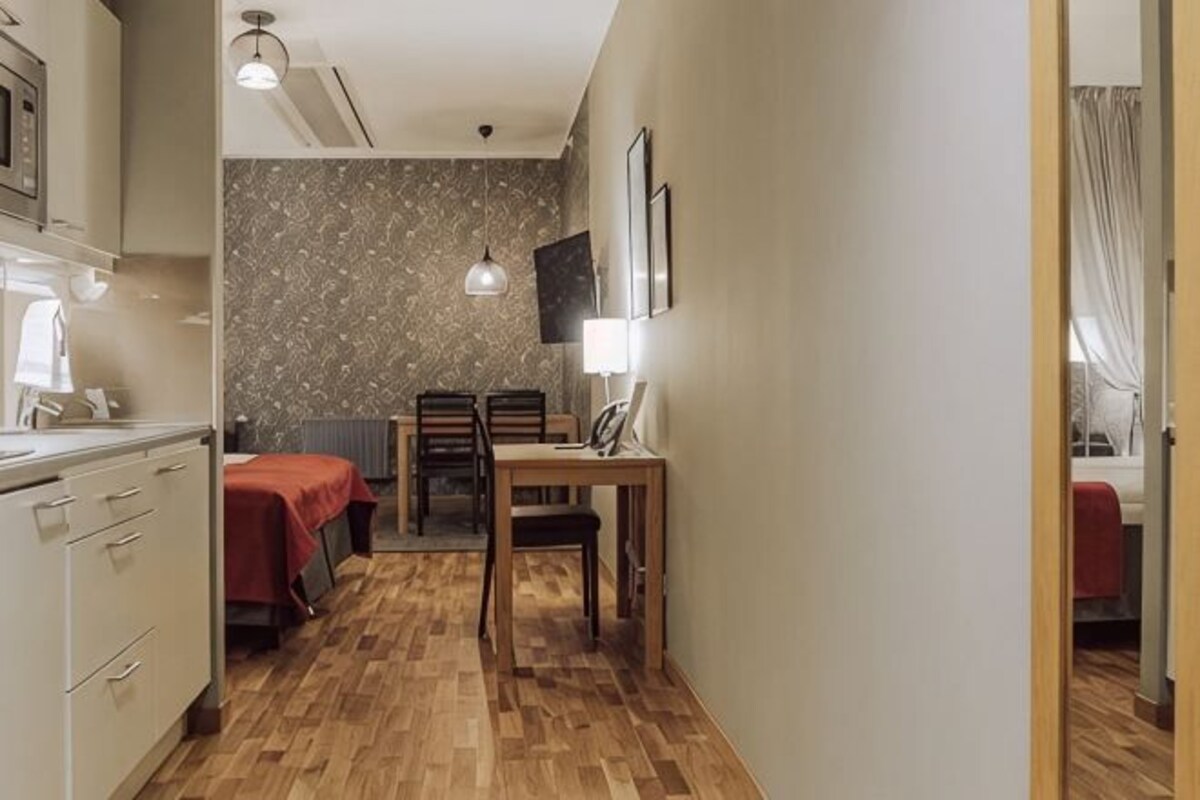 Habitación de hotel compacta con piso de madera, cama, escritorio con silla, una pequeña mesa con lámpara y televisor en la pared.