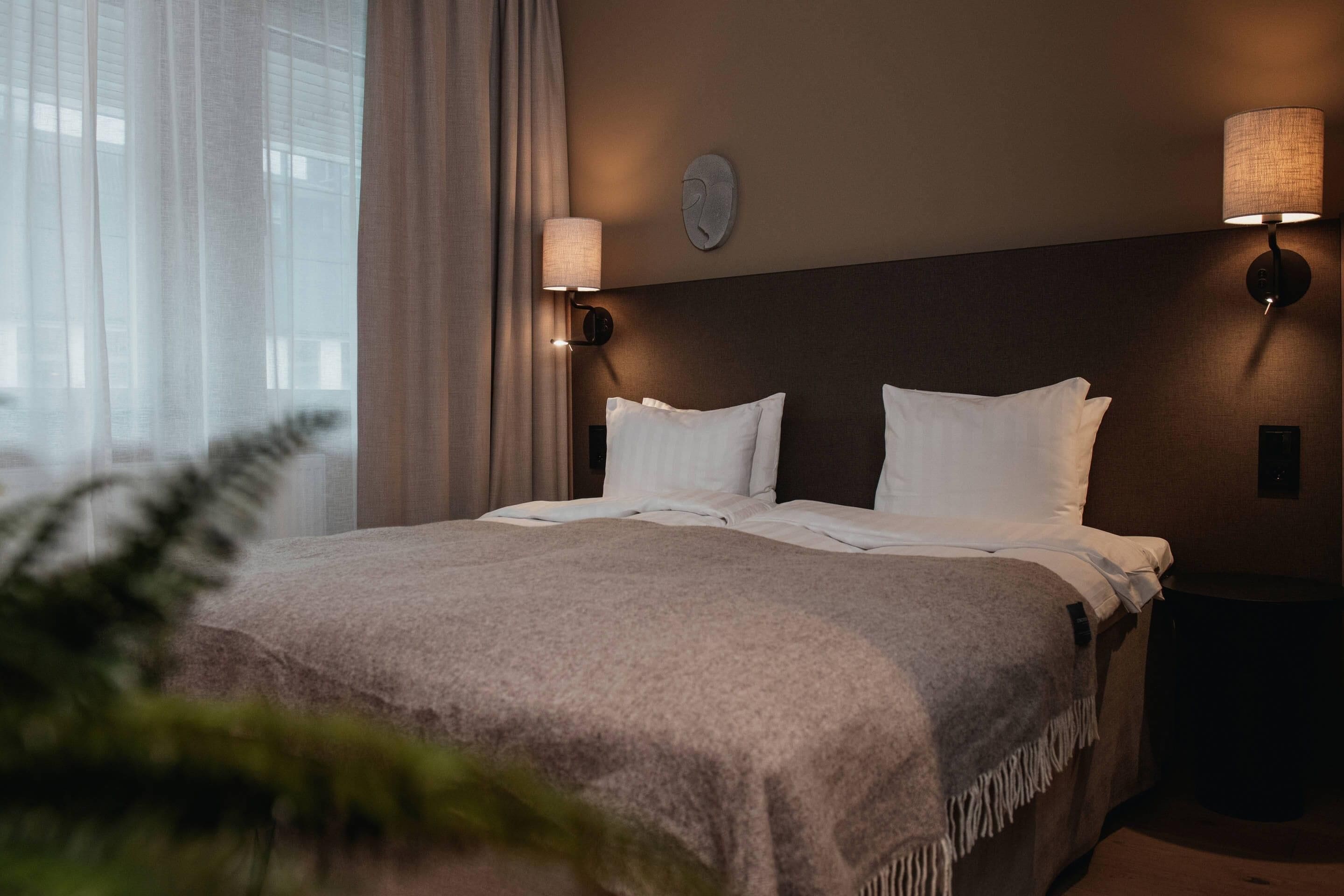 Een hotelkamer met een bed, twee wandlampen, een raam met gordijnen en een plant op de voorgrond.
