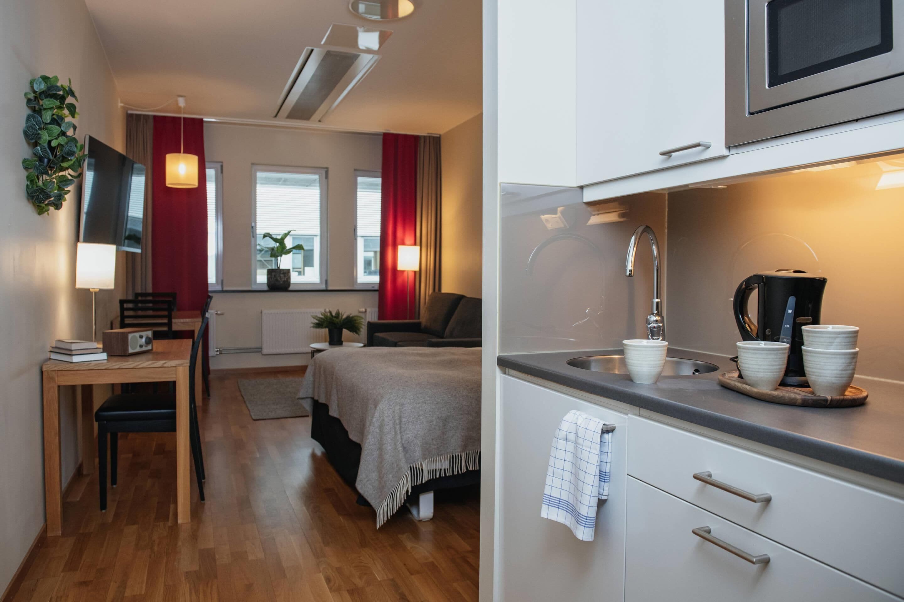 A modern room with a bed, a desk, a TV, and a kitchen area with a microwave and kettle.