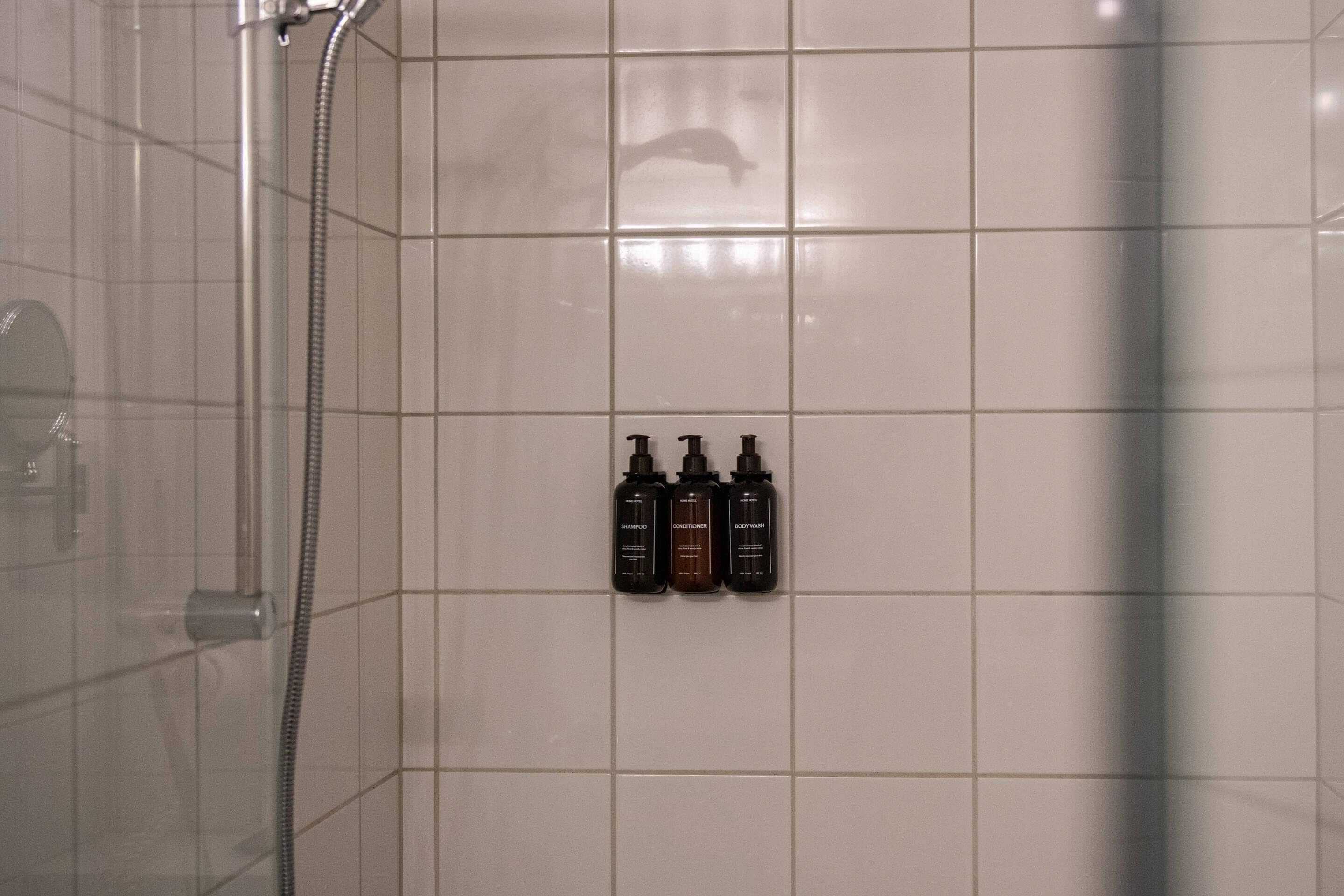 A tiled bathroom with a shower, a glass door, and three bottles of shampoo, conditioner, and body wash on the wall.
