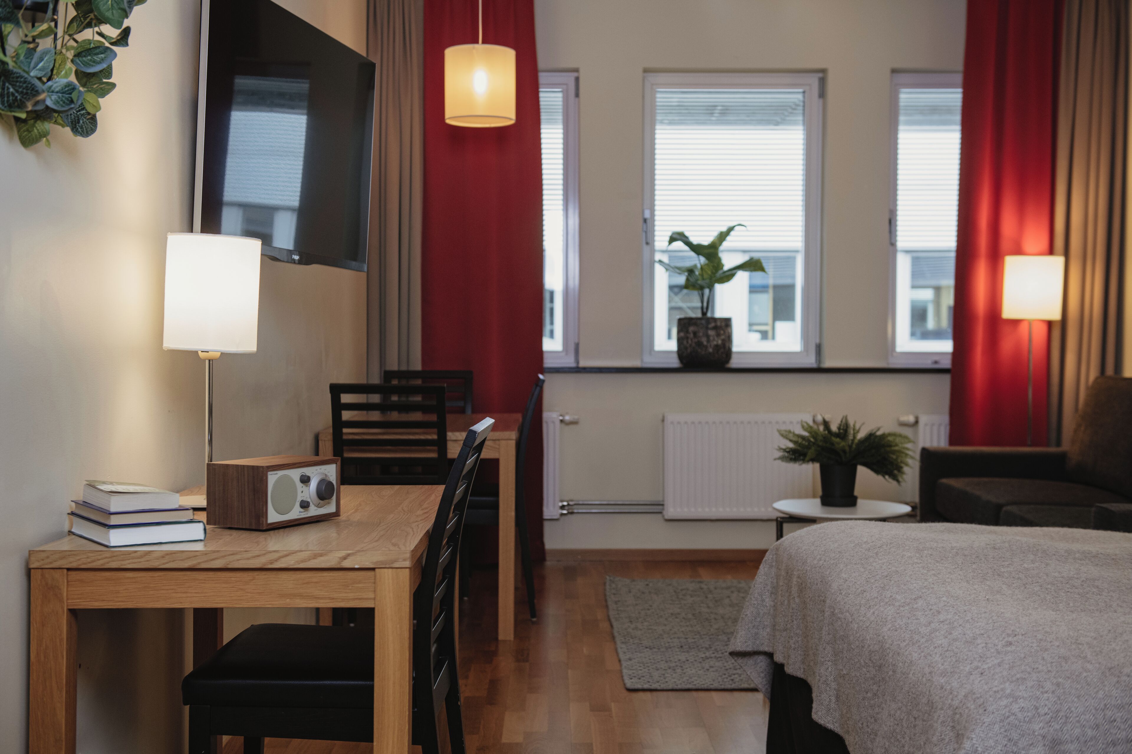 A modern hotel room with a wooden desk, a chair, a bed, a small table, and a potted plant.