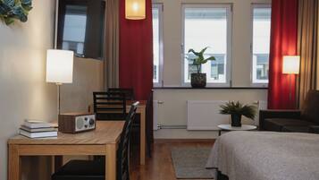 A modern hotel room with a wooden desk, a chair, a bed, a small table, and a potted plant.