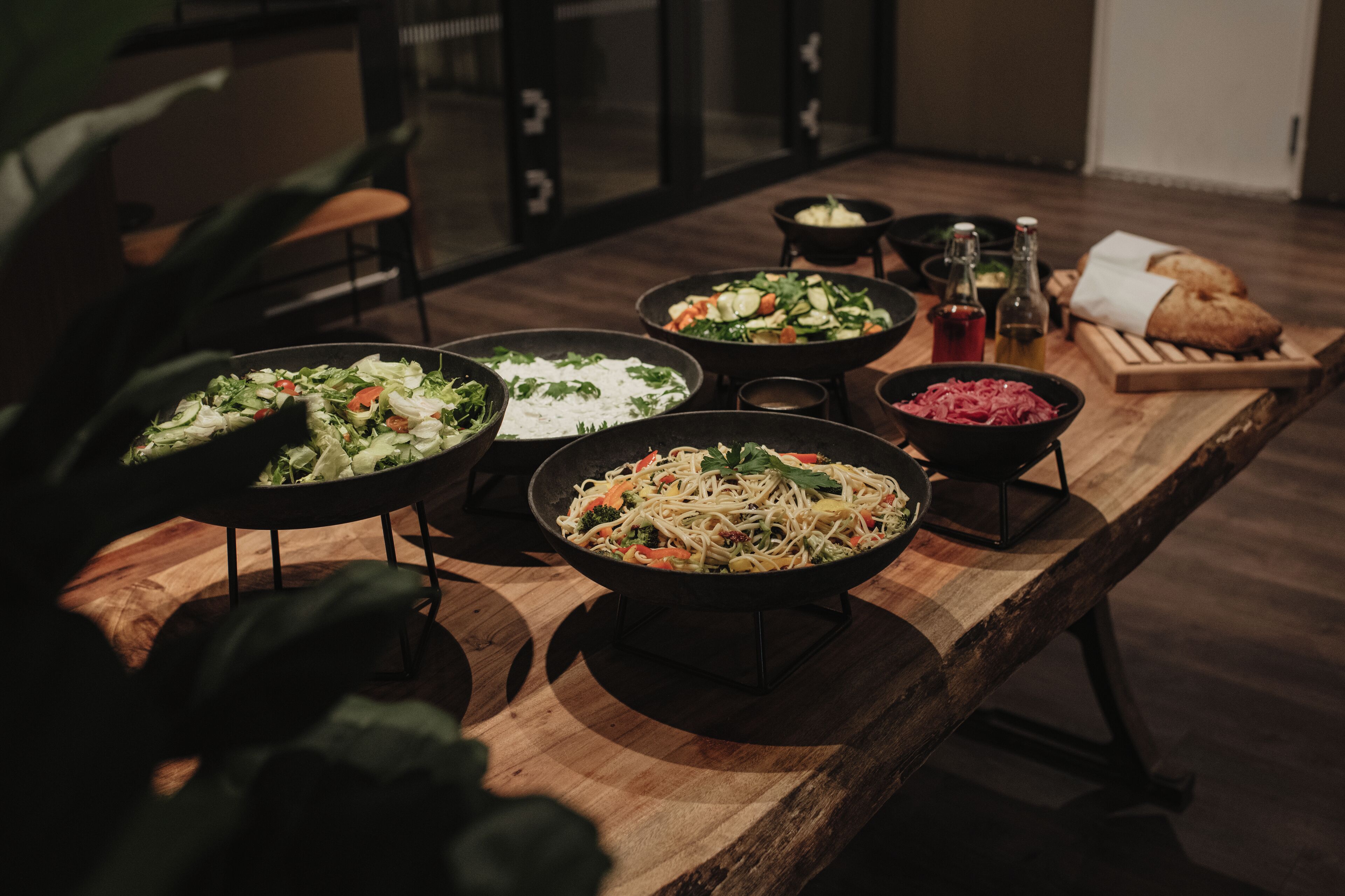 Una mesa de madera con varios platos, incluyendo pasta, ensaladas y pan, en una habitación con poca luz, con una planta y sillas al fondo.