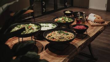 Una mesa de madera con varios platos, incluyendo pasta, ensaladas y pan, en una habitación con poca luz, con una planta y sillas al fondo.