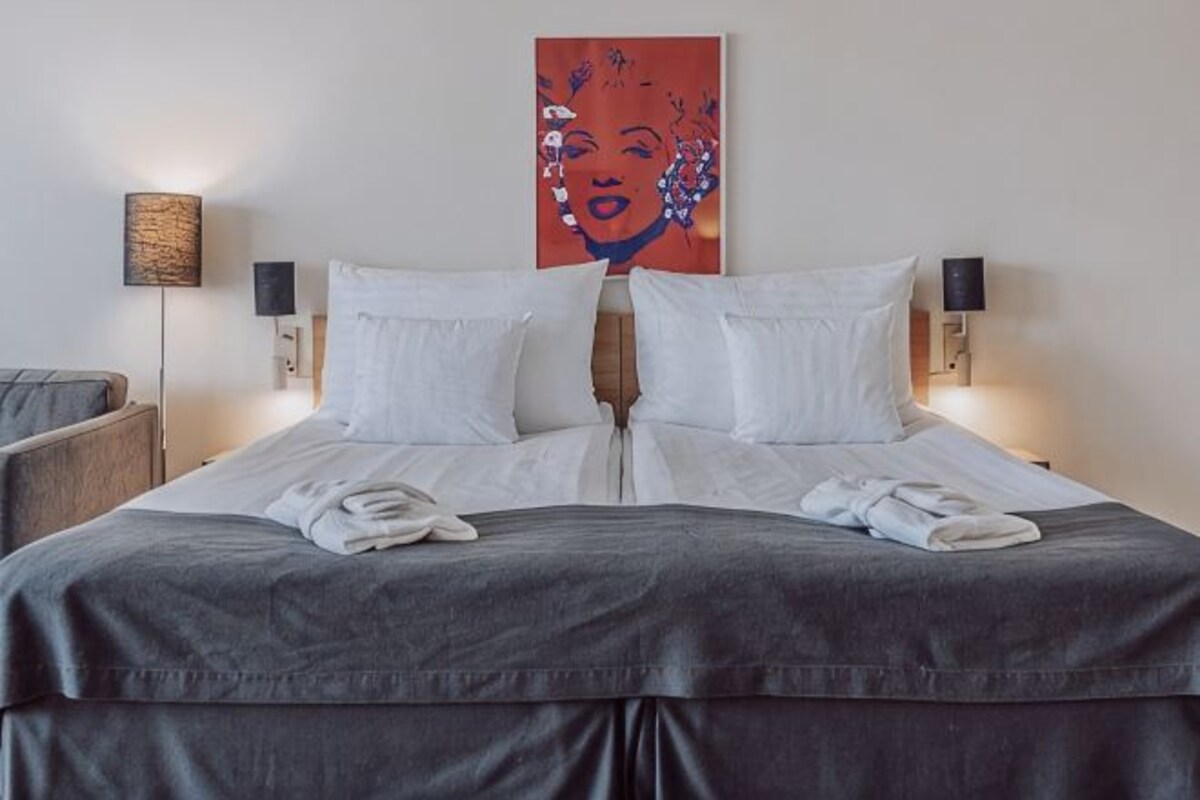 A neatly made bed with white linens, two wall-mounted lamps, and a large painting above the bed.